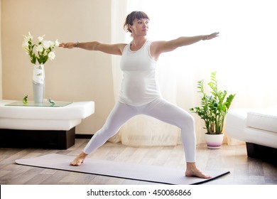 Healthy Pregnancy Yoga and Fitness concept. Young pregnant yoga woman working out in cozy living room interior. Pregnant model doing prenatal warrior II, virabhadrasana two yoga pose