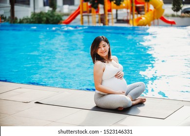 Healthy Pregnant Asian Chinese Woman Doing Yoga And Exercising Outdoor Near Swimming Pool. Lady With White Top And Tight Grey Pants. Relaxing Exercise. Cheerful Expecting Mother. Healthy Lifestyle.