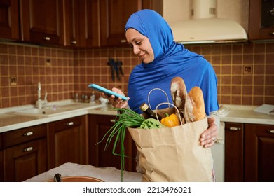 Healthy purchasing from grocery. Food delivery. Beautiful Arab Muslim woman in blue hijab, checking expenses while arriving home after grocery shopping and unpacking a shopping bag with healthy food - Powered by Shutterstock