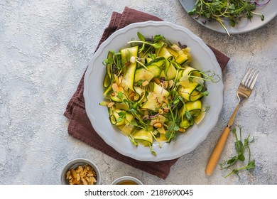 A healthy salad of raw zucchini, cheese, a mixture of microgreens and roasted almond slices with mustard dressing on a gray ceramic plate on a light concrete background. Salad recipes. Healthy food