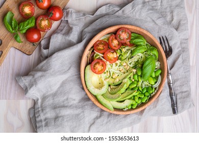 A healthy salad with zoodles zucchini noodles, baby tomatoes, avocado and edamame beans. The fresh salad is in a wooden bowl on a white table, with a linen napkin underneath the bowl.