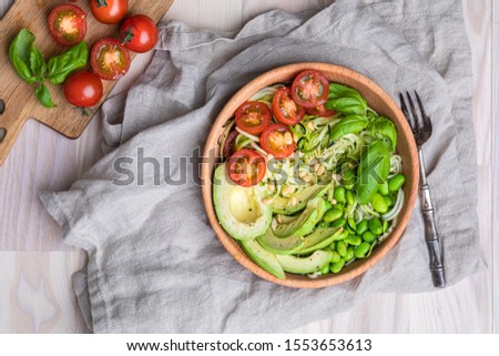 A healthy salad with zoodles zucchini noodles, baby tomatoes, avocado and edamame beans. The fresh salad is in a wooden bowl on a white table, with a linen napkin underneath the bowl. Stock photo © 