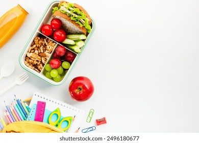 Healthy school lunch with sandwich, fruits, and veggies on a white desk with school supplies. Perfect for back-to-school or office lunch themes