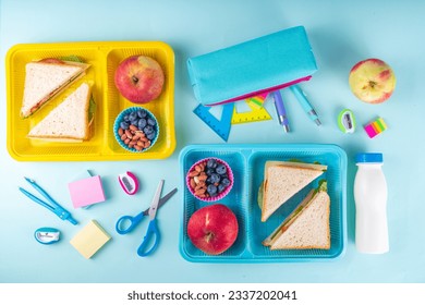 Healthy school meal, children packed lunch box with fruit, berry, nuts and sandwich with vegetables. Kids diet snack food with education school supplies, bright blue background top view copy space