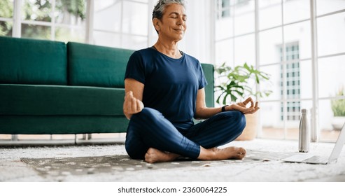 Healthy senior woman meditating in lotus position as she follows an online fitness class. Mature woman practicing yoga in front of a laptop at home, finding relaxation and mindfulness in retirement. - Powered by Shutterstock