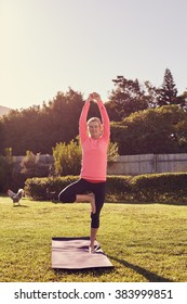 Healthy Senior Woman On Her Yoga Mat Outdoors On The Grass In Her Garden, With Summer Morning Sunflare And Clear Sky Above Her