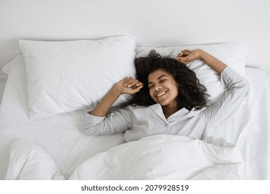 Healthy Sleeping Concept. Above View Of Happy Dark Skinned Woman Waking Up In White Bed, Raised Hands Up, Stretch Body And Smiling Wide. Female Awaking And Spending Lazy Morning In Pajamas