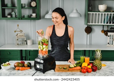 Healthy slim girl adding cucumber to the blender full of fresh vegetables for making healthy drink in the kitchen. Smoothie and super food eating for healthy diet and body slimming - Powered by Shutterstock