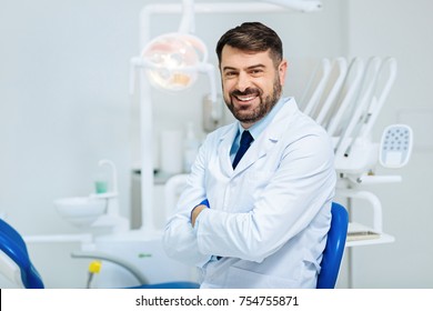 Healthy smile. Positive bearded dentist expressing cheer while standing in front of you and keeping arms in crossed position - Powered by Shutterstock