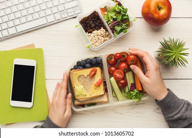 Healthy snack at office workplace. Businesswoman eating organic vegan meals from take away lunch box at wooden working table with computer keyboard and smartphone