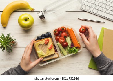 Healthy snack at office workplace. Businesswoman eating organic vegan meals from take away lunch box at wooden working table with computer keyboard, top view