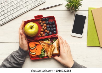 Healthy snack at office workplace. Businesswoman eating organic vegan meals from take away lunch box at wooden working table with computer keyboard and smartphone with empty screen for copy space