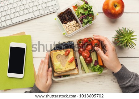 Healthy snack at office workplace. Businesswoman eating organic vegan meals from take away lunch box at wooden working table with computer keyboard and smartphone