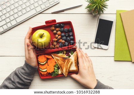 Healthy snack at office workplace. Businesswoman eating organic vegan meals from take away lunch box at wooden working table with computer keyboard and smartphone with empty screen for copy space