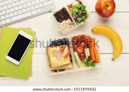 Healthy snack at office workplace. Eating organic vegan meals from take away lunch box at wooden working table with computer keyboard and smartphone with empty screen for copy space, top view