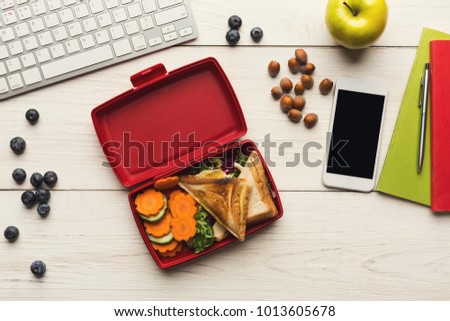 Healthy snack at office workplace. Eating organic vegan meals from take away lunch box at wooden working table with computer keyboard and smartphone with empty screen for copy space, top view