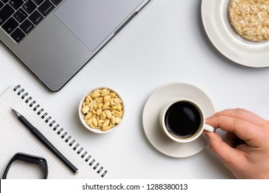 Healthy snacking at work during break time. Sportsman eating crispy rice rounds with peanuts, cup of coffee near the laptop, fitness-tracker and notebook. White organized desk. - Powered by Shutterstock