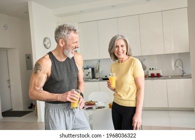 Healthy sporty happy senior adults family couple drinking juice and laughing at home. Cheerful fit mid age older husband and wife enjoying talking, having fun standing in kitchen in the morning.