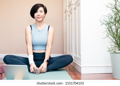 Healthy and stylish Asian woman sit in easy seat pose or sukhasana practicing yoga online class via computer tablet, look at camera and smile in bedroom at home. Wellbeing and active lifestyle.