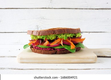 Healthy Superfood Sandwich With Avocado, Beet Hummus, Spinach Radishes And Carrots. Side View On A Wooden Board Against A White Wood Background.