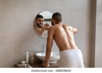 Healthy Teeth, Oral Care Concept. Rear Back View Of Smiling Muscular Bearded Guy In White Towel Brushing Teeth With Paste Looking In Round Mirror In Modern Interior Bathroom At Home Or Hotel Room