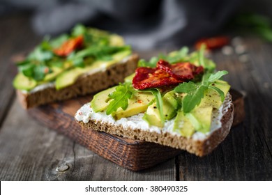 Healthy toast with goats cheese, avocado, arugula and sun dried tomatoes on rustic wooden table. Selective focus, close up