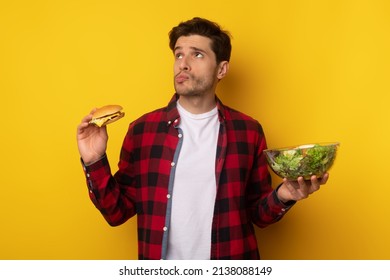 Healthy Or Unhealthy Food Concept. Funny Pensive Man Holding Bowl With Salad And Burger In Hand. Hungry Guy Choosing What To Eat, Thinking And Looking Up Isolated Over Orange Studio Background