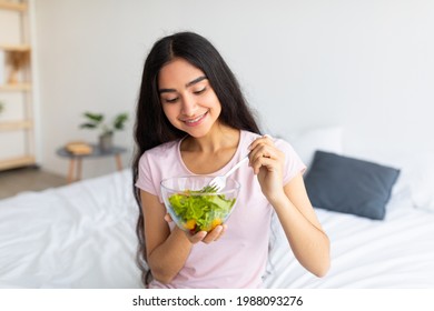 Healthy vegan diet concept. Joyful Indian lady sitting on bed at home with bowl of fresh vegetable salad, eating tasty lunch. Pretty Eastern woman enjoying her vegan meal, promoting clean living