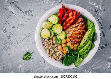 Healthy vegetable buddha bowl lunch with grilled chicken and quinoa, spinach, avocado, brussels sprouts, red paprika and chickpea on gray background. Top view.