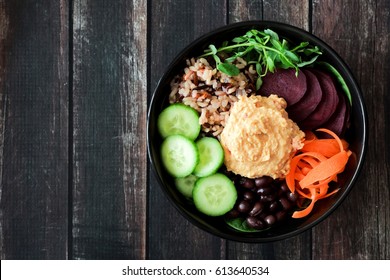 Healthy vegetarian nourishment bowl with hummus, beans, wild rice, beets, carrots, cucumbers and pea shoots. Above view on dark wood.