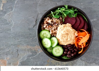 Healthy vegetarian salad bowl with hummus, beans, wild rice, beets, carrots, cucumbers and pea shoots. Above view on slate background. 