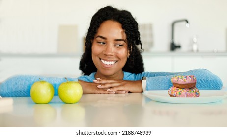 Healthy Vs Unhealthy Diet. Black Woman Looking At Donuts And Apples, Making Choice Of Her Meal. Sporty Lady Choosing Between Balanced And Unbalanced Nutriton