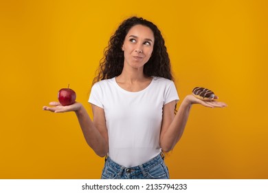 Healthy Vs Unhealthy Food Concept. Puzzled Pensive Funny Woman Holding Donut And Red Apple Fruit. Hungry Latin Lady Choosing What To Eat, Thinking And Posing Isolated On Yellow Orange Studio Wall