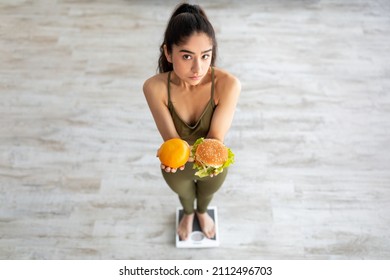 Healthy Vs Unhealthy Nutrition. Above View Of Young Indian Woman Holding Citrus Fruit And Hamburger, Standing On Scales, Making Choice. Millennial Lady Choosing Between Balanced And Unbalanced Diet