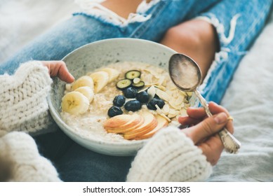 Healthy Winter Breakfast In Bed. Woman In Woolen Sweater And Shabby Jeans Eating Vegan Almond Milk Oatmeal Porridge In Bowl With Berries, Fruit And Almonds. Clean Eating, Vegetarian Food Concept