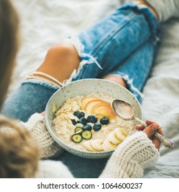 Healthy Winter Breakfast In Bed. Woman In Woolen Sweater And Shabby Jeans Eating Vegan Almond Milk Oatmeal Porridge With Berries, Fruit And Almonds, Square Crop. Clean Eating, Vegetarian Food Concept