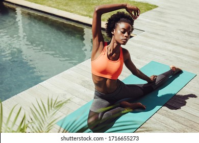 Healthy woman doing yoga stretches by the poolside. African female in sportswear exercising yoga in morning by the swimming pool - Powered by Shutterstock