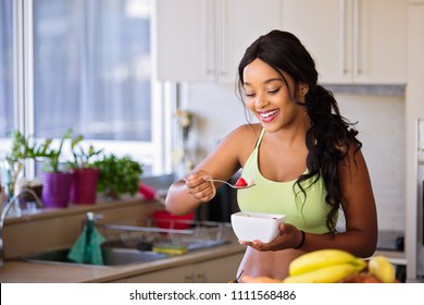Healthy woman eats from a bowl of fruit with strawberries. She is dressed in sports wear ready for a workout. Very happy and lean with toned body. In kitchen. - Shutterstock ID 1111568486