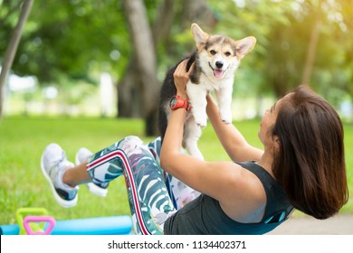 A healthy woman playing with a corgi puppy while excersing on yoga mat surrounding with gym tools such as kettlebell and dumbbell, outdoor training with dog
