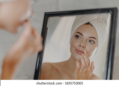 Healthy woman wears minimal makeup, takes care of complexion and lips, looks at herself in mirror, stands bare shoulders, wears bath towel on head, has healthy flawless skin. Beauty, hygiene concept