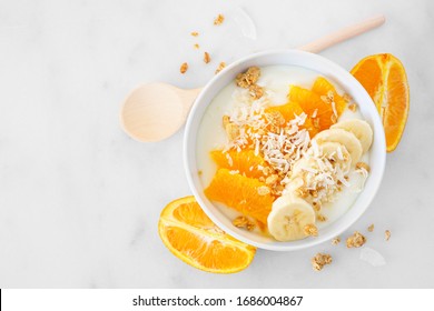 Healthy Yogurt Bowl With Oranges, Banana And Coconut. Overhead View Table Scene On A White Marble Background.