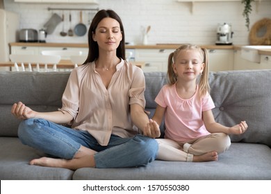 Healthy young adult parent mother and little child daughter doing yoga exercise sit on sofa at home, calm mom teaching cute small kid girl learning practice family meditation relax together concept