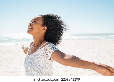 Healthy young african woman with curly hair standing at beach with hands outstretched and looking up. Happy young black woman with open arms feeling the sea breeze with copy space. Beautiful girl. - Powered by Shutterstock