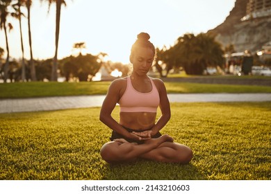 Healthy young African woman in sportswear meditating in the lotus position while sitting alone in a park practicing yoga  - Shutterstock ID 2143210603