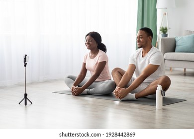 Healthy young black couple exercising on yoga mat at home, watching online fitness lesson or having video call with yoga trainer, using smartphone on tripod, panorama with copy space
