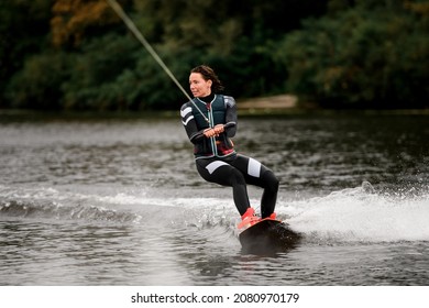 Healthy young female wakeboarder in wetsuit and vest riding on wakeboard on river water at summer day