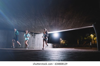 Healthy Young People Running Under A Bridge At Night. Group Of Runners Training In The City.