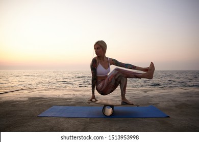 Healthy Young Pretty Tattooed Woman With Casual Hairstyle Making Exercises On Sports Equipment Outdoor, Posing Over Sea View On Balance Board, Balancing On Wooden Desk During Sunrise