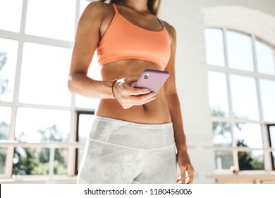 Healthy Young Woman Counts Calories And Made Post In Blog After Fitness Workout. Sporty Tanned Girl In White Sunny Gym Hold Mobile Phone In Hand. Sport And Gadget Concept