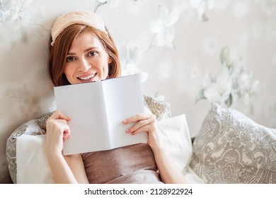 A healthy young woman wearing a sleeping mask after a good night's rest. A happy red-haired lady feels cheerful, full of joy, full of energy in the early morning, reading a book. Crazy home